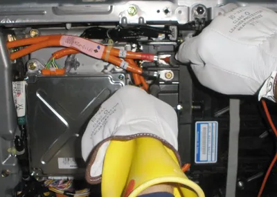 A technician wearing insulated safety gloves working on the bright orange high-voltage cables of a hybrid vehicle.